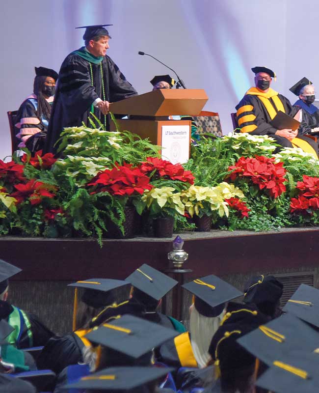 Speaker adorned in cap and gown on stage at podium with others seated behind him, and graduates adorned in cap and gown seated in audience in front of him