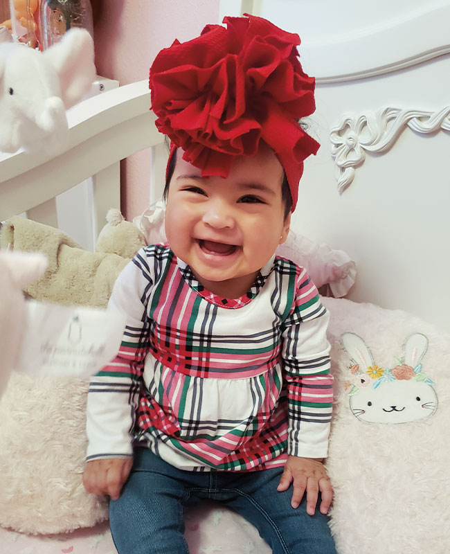 Ariel Chavez baby in white crib wearing red bow on head and plaid shirt