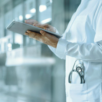Torso of a person in white lab coat with stethoscope in pocket and holding a tablet device