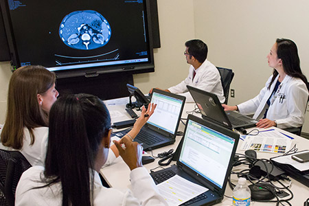 Doctors sitting at a meeting room table with laptops in front of them and a flatscreen on the wall displaying a clinical image