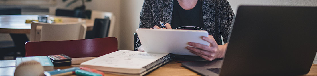 woman sits at desk taking notes on paper and tablet near laptop