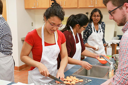 3 women 1 man in aprons prepare food in kitchen