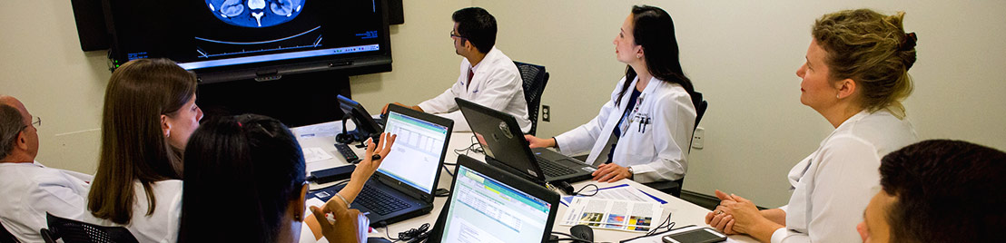 Doctors sitting at a meeting room table with laptops in front of them and a flatscreen on the wall displaying a clinical image
