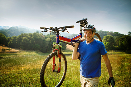 Man in a bike helmet and a blue shirt carries a red bicycle on his shoulder in a field