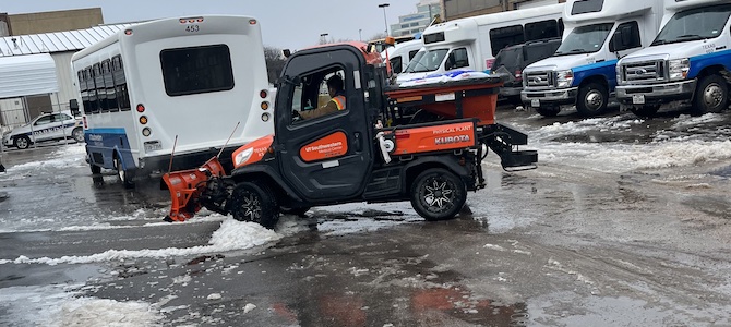 An orange snow plow clears a parking lot where UTSW shuttle buses are parked