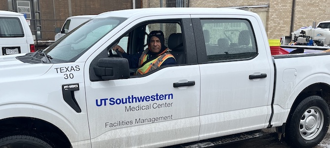 A male UTSW worker wears a dark cap and an orange and yellow safety vet as he sits in a white pickup truck